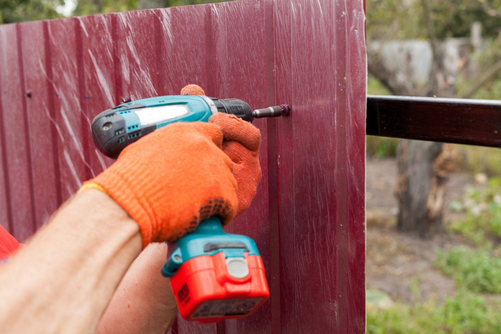 Person wearing orange gloves using a teal and black drill to attach a burgundy metal panel to a fence.
