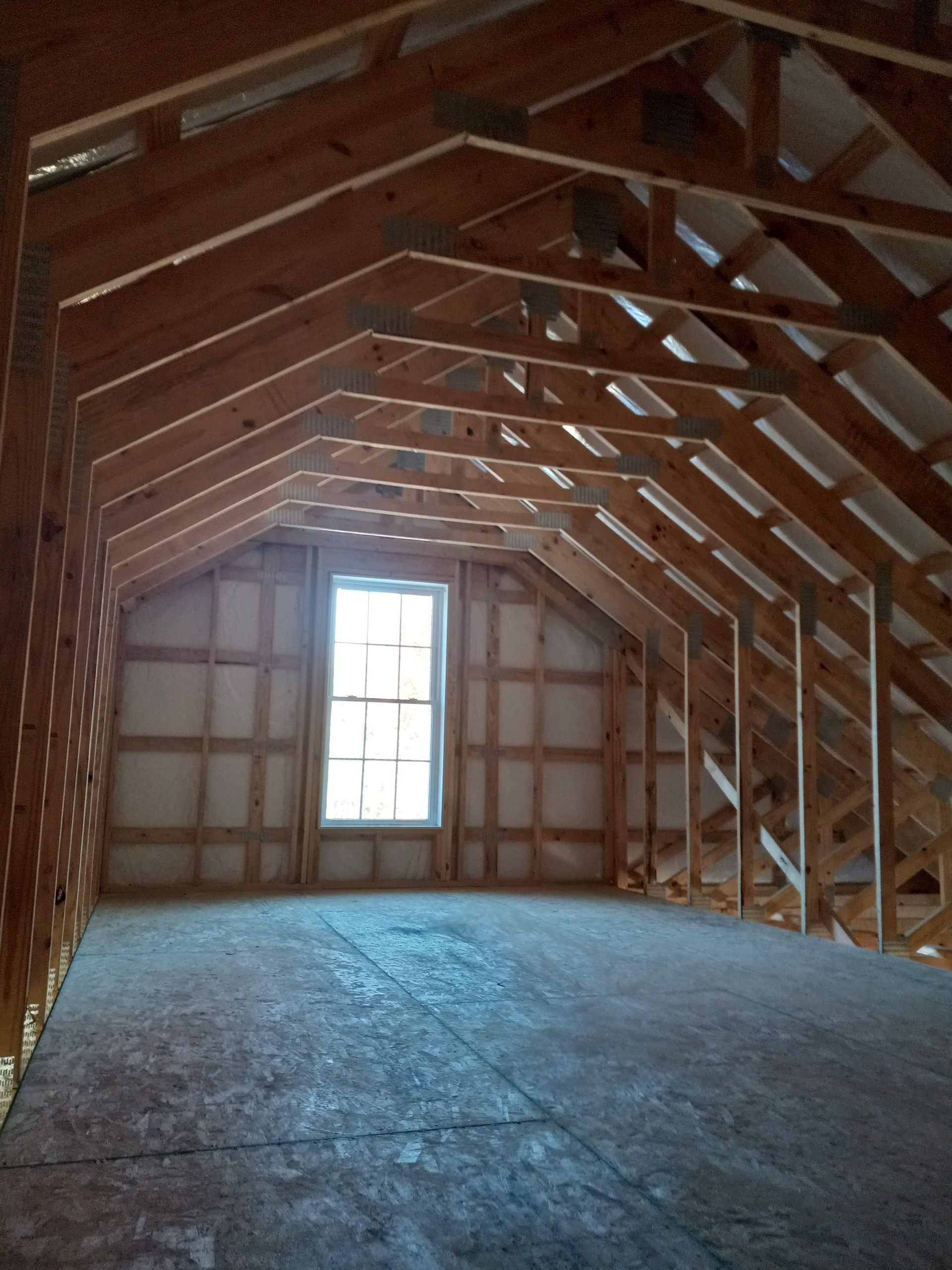 Empty unfinished attic with exposed wooden roof trusses and a small window at the far end.