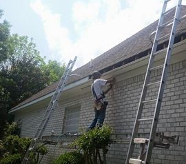 A construction worker on a scaffolding platform repairs the fascia board on a brick house under a blue sky.