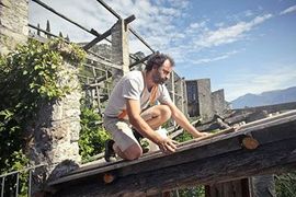 A person kneeling on a wooden roof structure, installing planks against a backdrop of stone ruins and mountains.