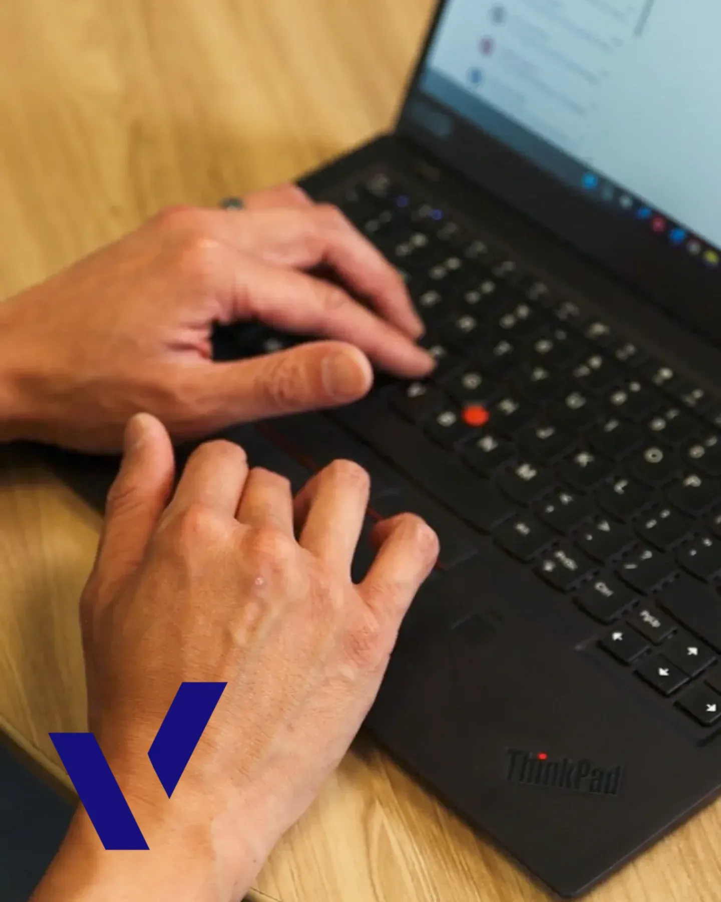 Hands typing on a black ThinkPad laptop keyboard atop a wooden desk surface.