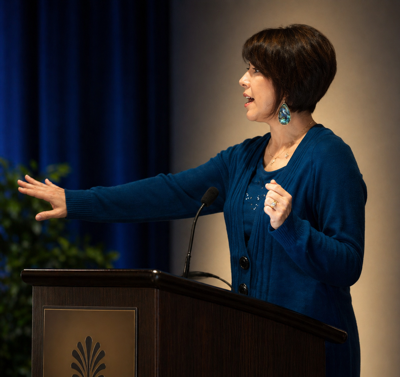 Woman speaking at podium, gesturing with outstretched arm on a stage with blue lighting