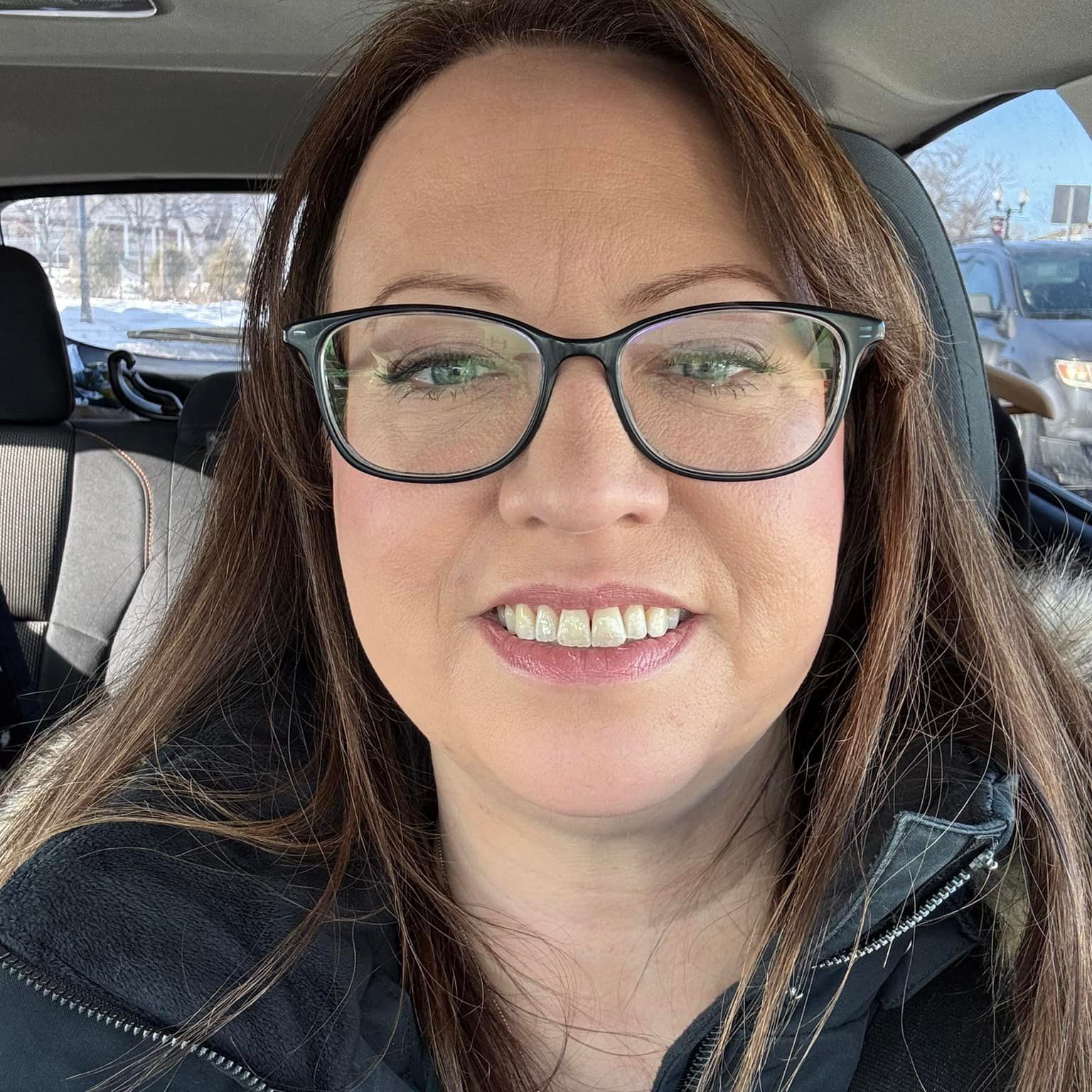 Smiling woman with glasses in a car, wearing a black jacket and scarf, snowy street visible outside.
