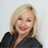 Smiling blonde woman in a dark blazer against a light gray background