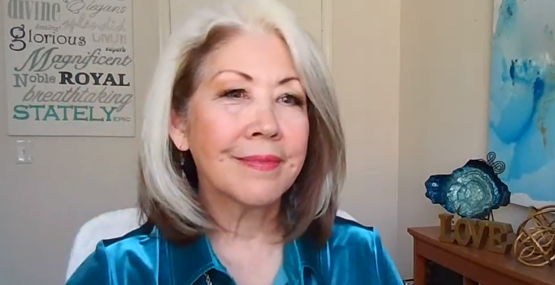 Smiling woman in a teal blouse in a home office, with wall art and a shelf behind her