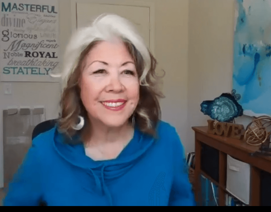 Smiling woman in a blue top sitting indoors, with wall art and a shelf in the background