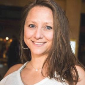Smiling woman with long brown hair in a white top, indoors with a softly blurred background