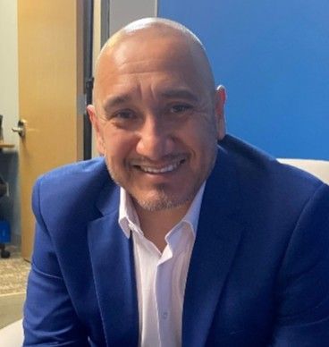 Smiling bald man in a blue blazer and white shirt seated indoors against a blue wall