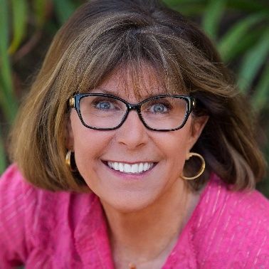 Smiling woman with glasses and gold hoops in a pink top, against a blurred outdoor background