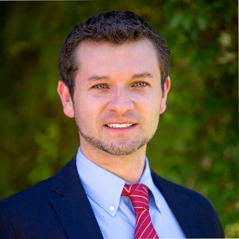 Man in a navy suit and red tie smiling outdoors against a blurred green background