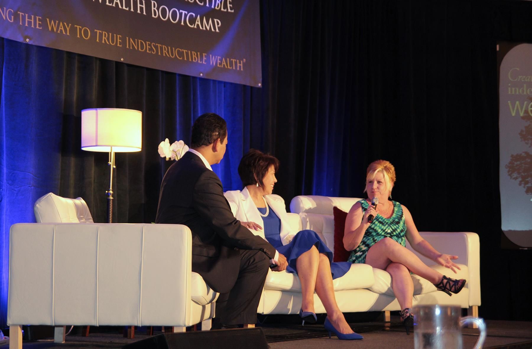 Three people sit on a stage in a panel discussion, with a lamp and banner in the background.