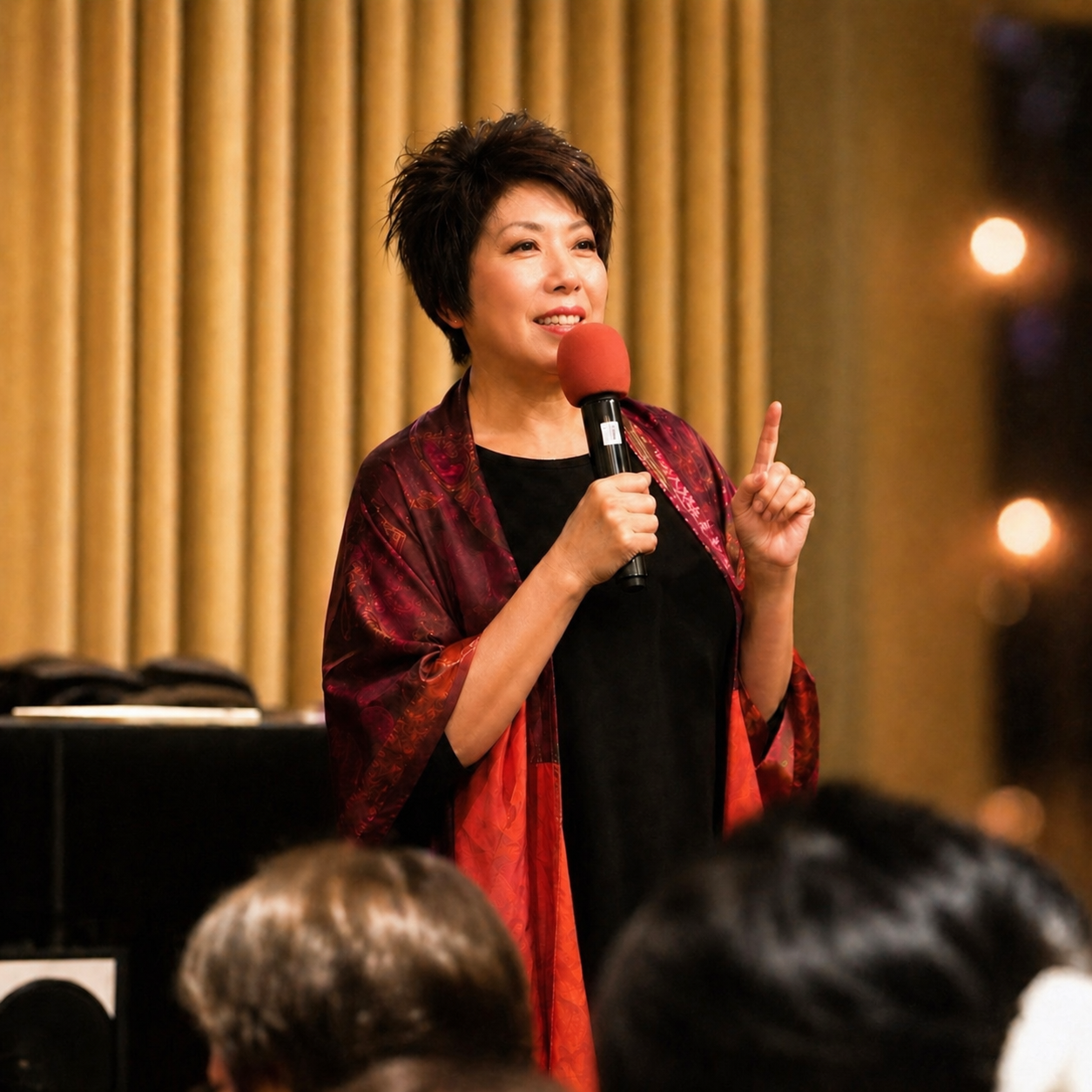 Speaker holding a red microphone on stage, wearing a black dress and red shawl, with audience in foreground