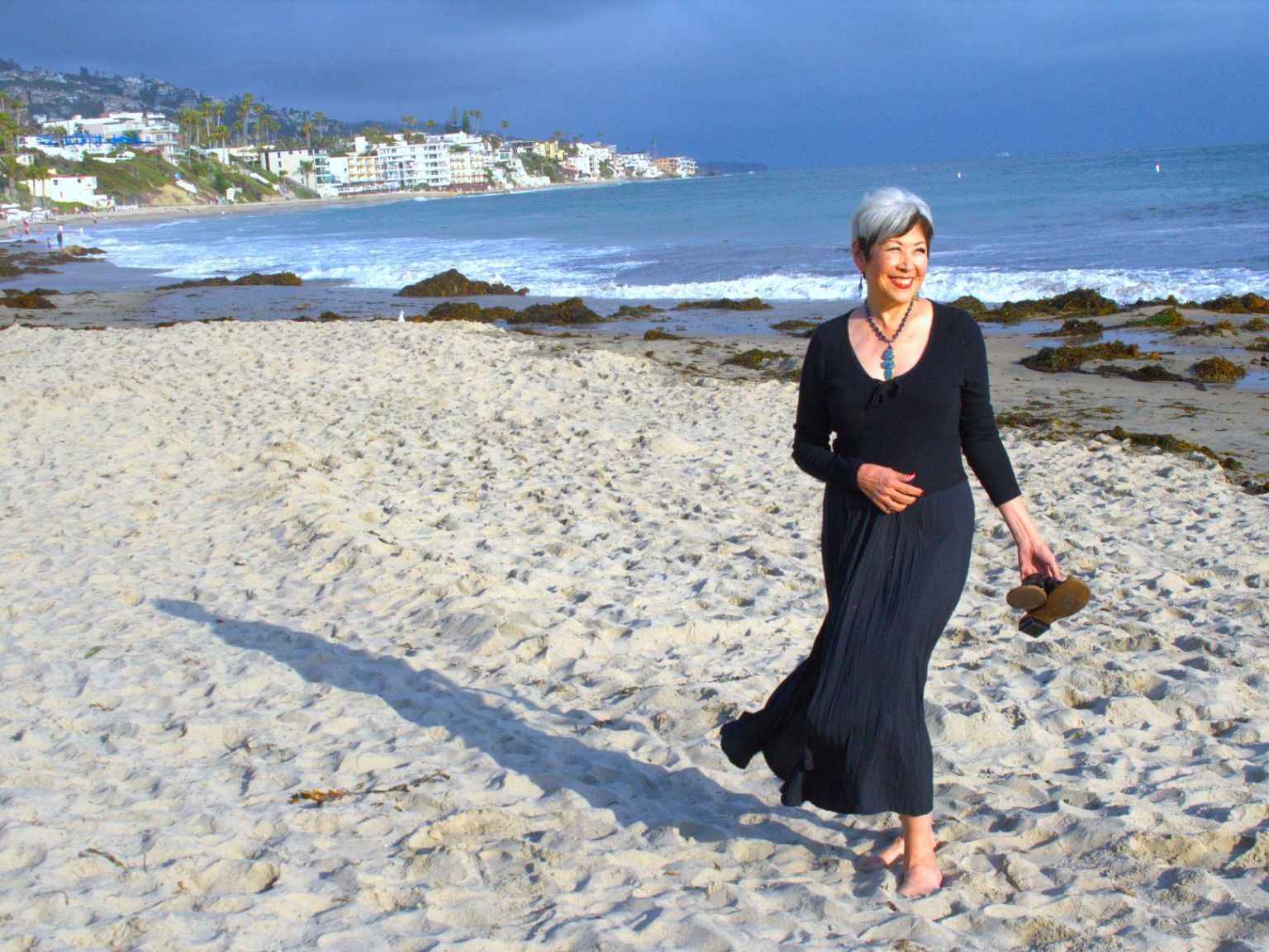 Woman in a black dress walking barefoot on a sunny beach near the shoreline.