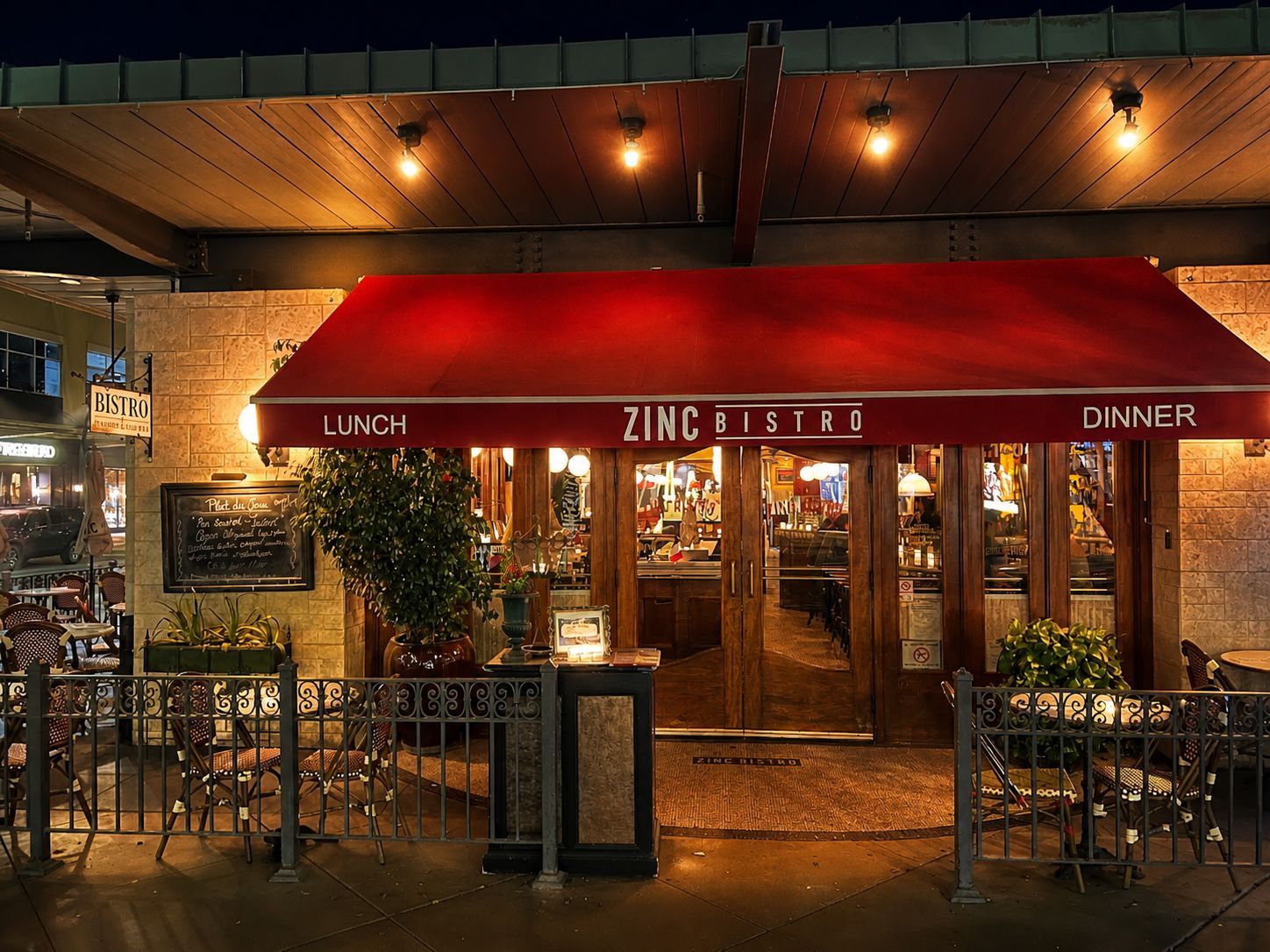 Restaurant entrance with red awning and warm lights at night, flanked by greenery and outdoor seating.