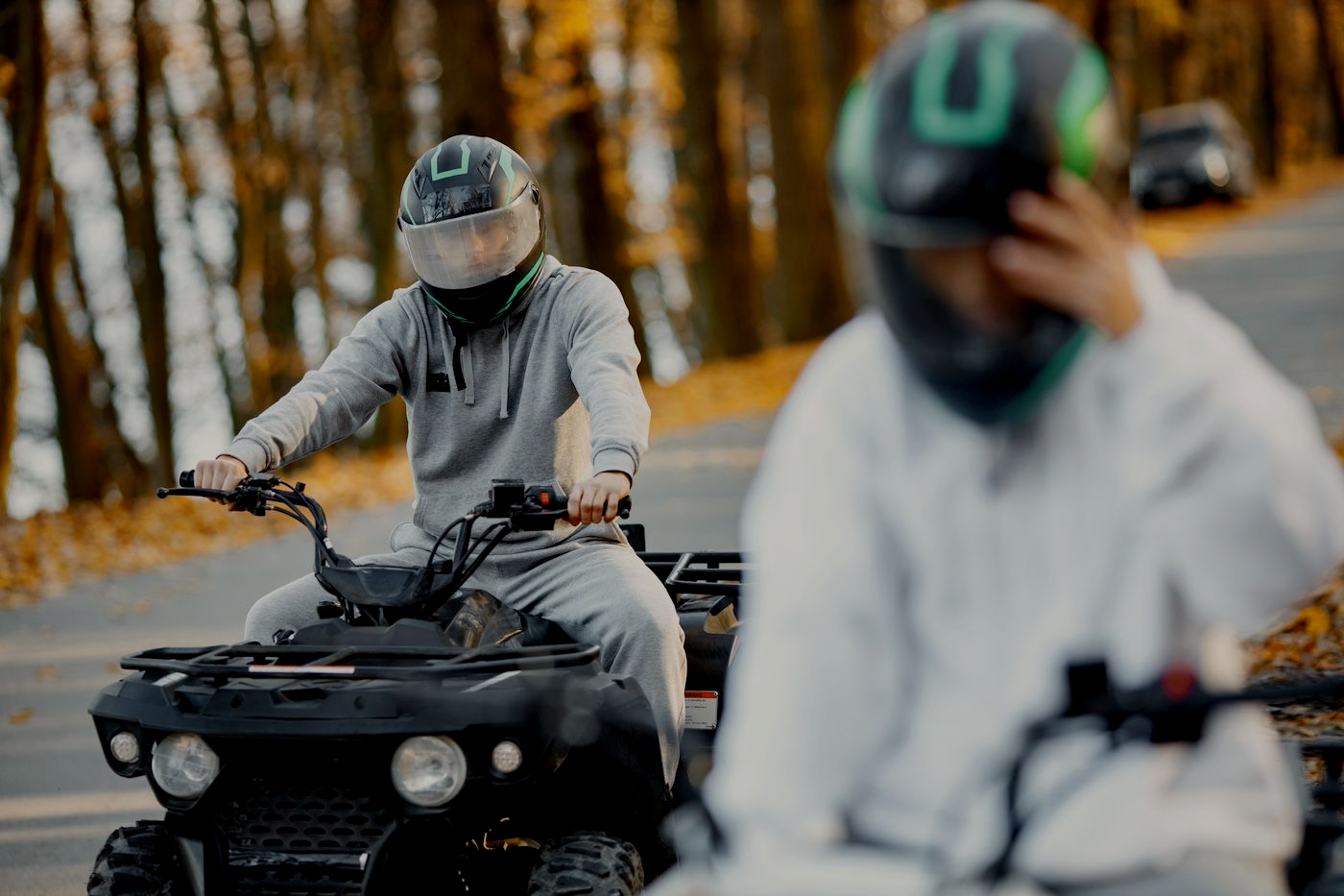 Two people ride ATVs on a wooded trail, one in focus wearing gray gear, the other blurred in front.