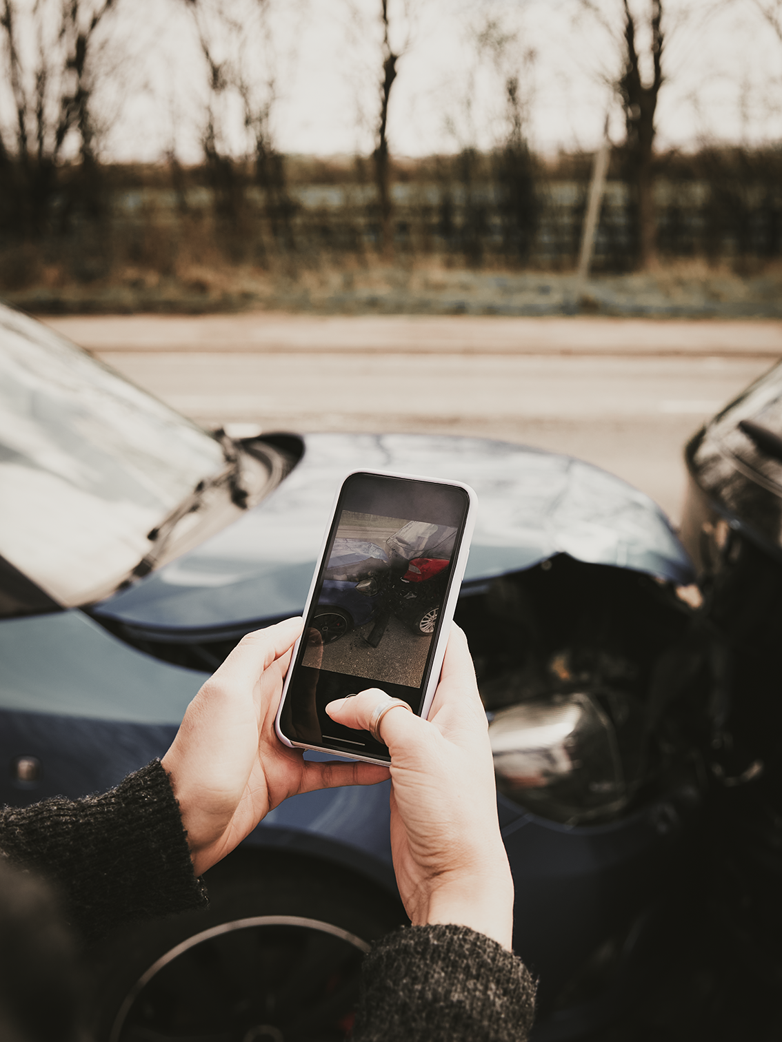 Hands holding a smartphone beside a car with a shattered windshield on a roadside