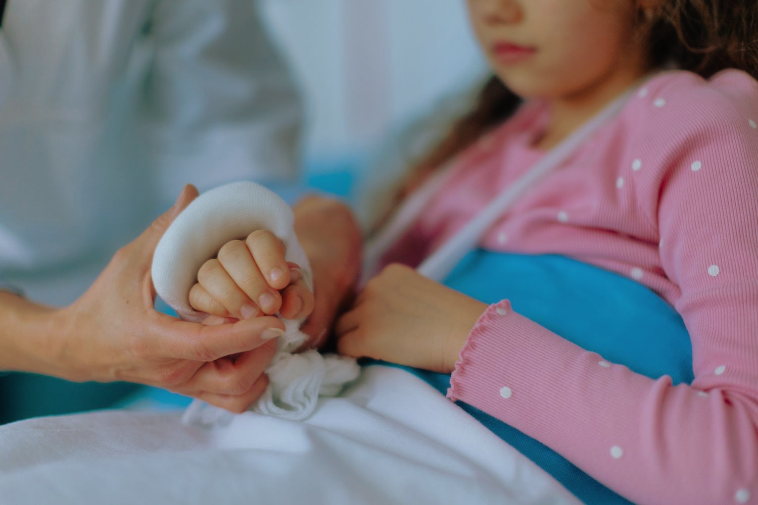 Adult hand comforting a child’s wrapped hand beside a patient in a pink sweater and blue arm sling