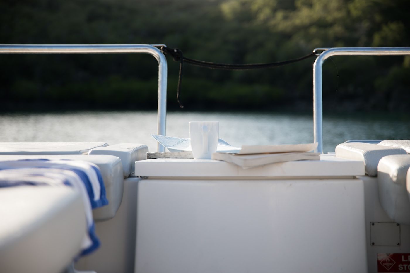 Back of a boat with a white surface, cup, book, and towel, overlooking a body of water and trees.