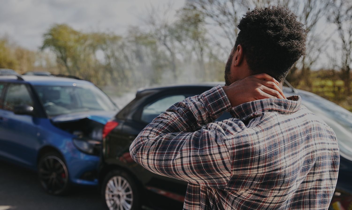 Person standing near two damaged cars on a roadside, holding the back of their neck.