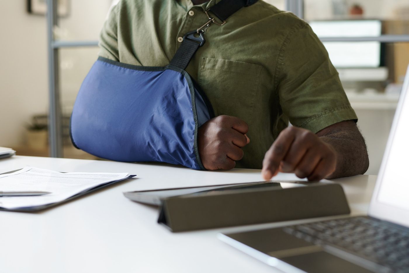 Person with arm in blue sling working at a desk with a laptop and papers