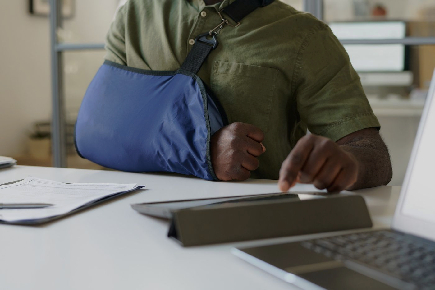 Person with arm in blue sling working at a desk with a laptop and papers