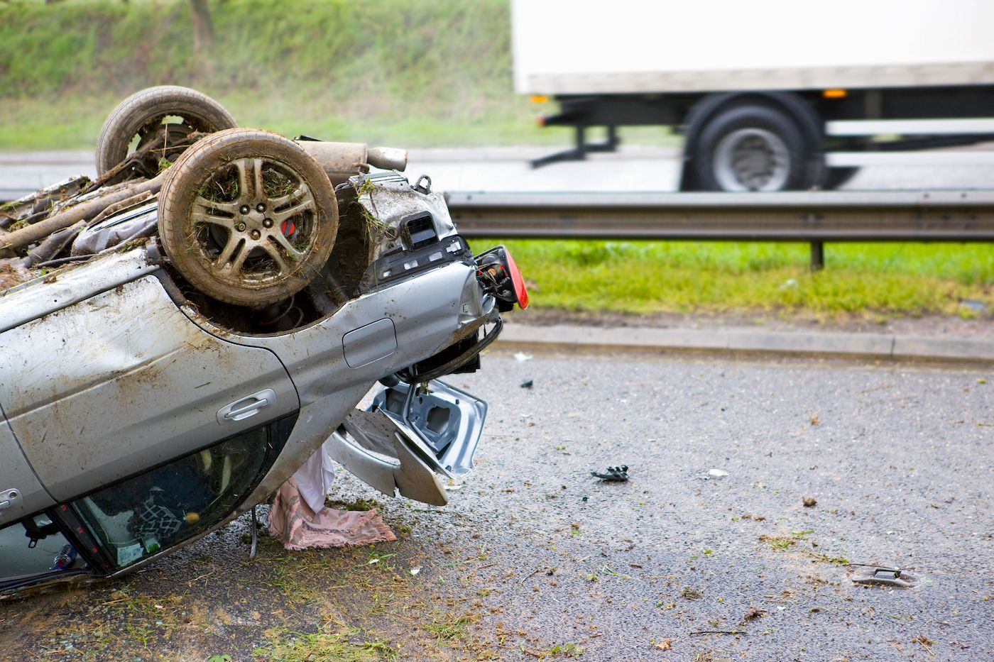 Overturned silver car on a wet road, with debris and a guardrail nearby after a crash