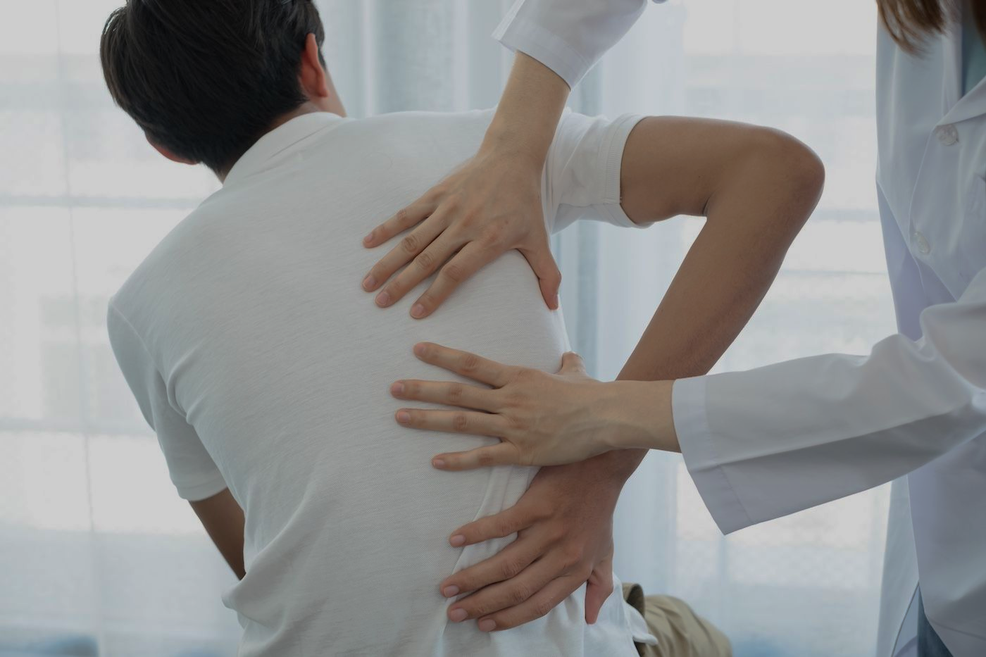 Doctor examining a patient’s back while another hand supports the spine in a bright clinic