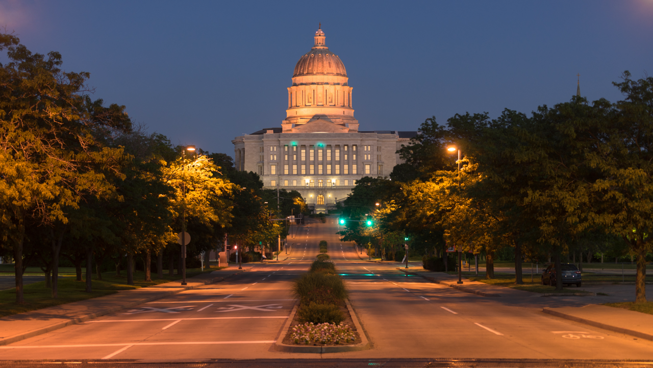 Illuminated capitol building at dusk, centered beyond a tree-lined avenue with empty streets