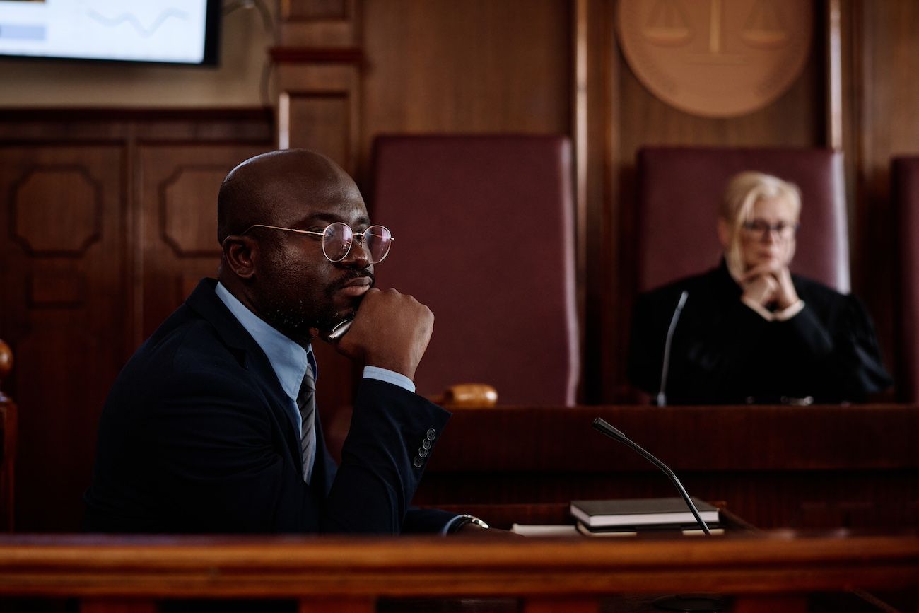 Man in suit at a courtroom table, looking thoughtful. Judge sits in the background.