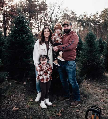 Family of four posing outdoors in a wooded area, wearing casual clothes and holding a young child. Family of four posing outdoors in a wooded area, wearing casual clothes and holding a young child.