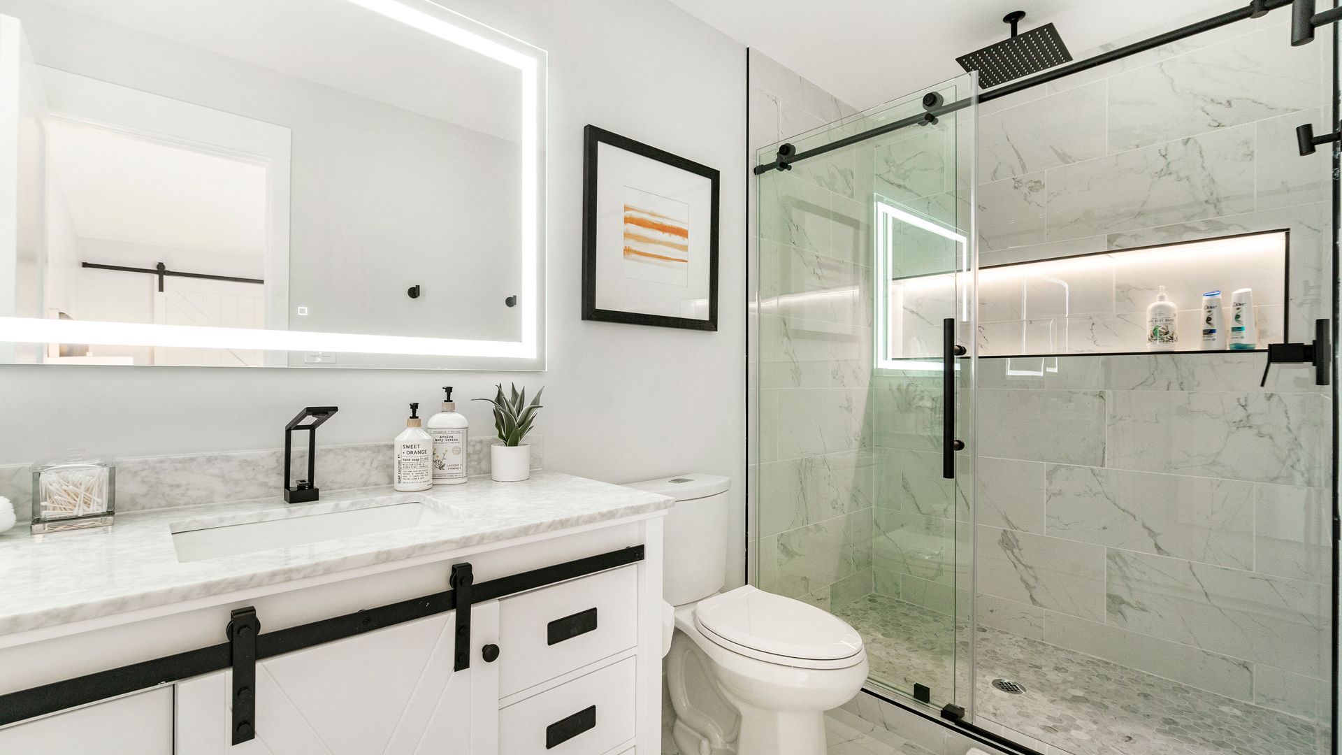 Modern bathroom with white vanity, black hardware, glass shower, lit mirror, and artwork.