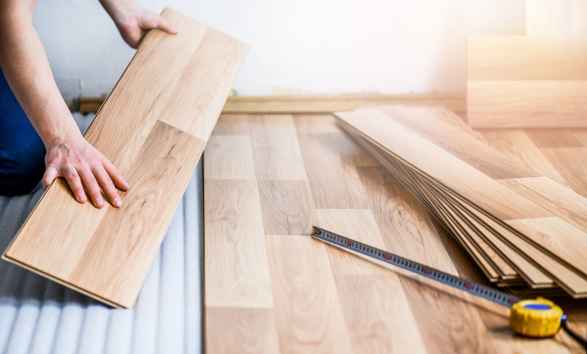 Person installing light brown laminate flooring; hands holding a plank, measuring tape visible.