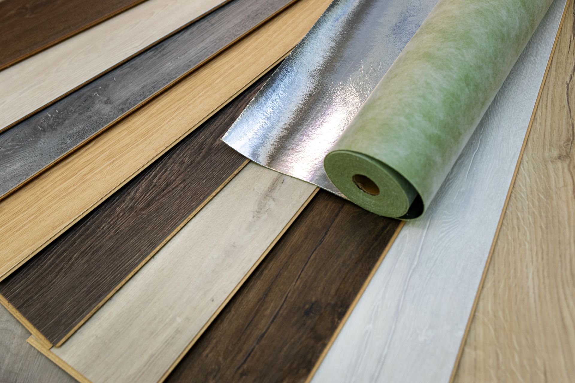 Assorted laminate flooring samples in various wood tones with an underlayment roll.
