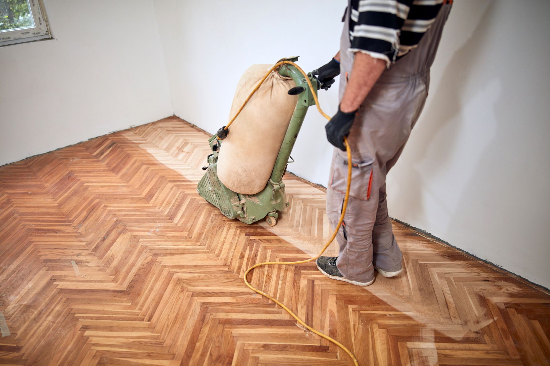 Person sanding wooden floor with a floor sander in a room with white walls.