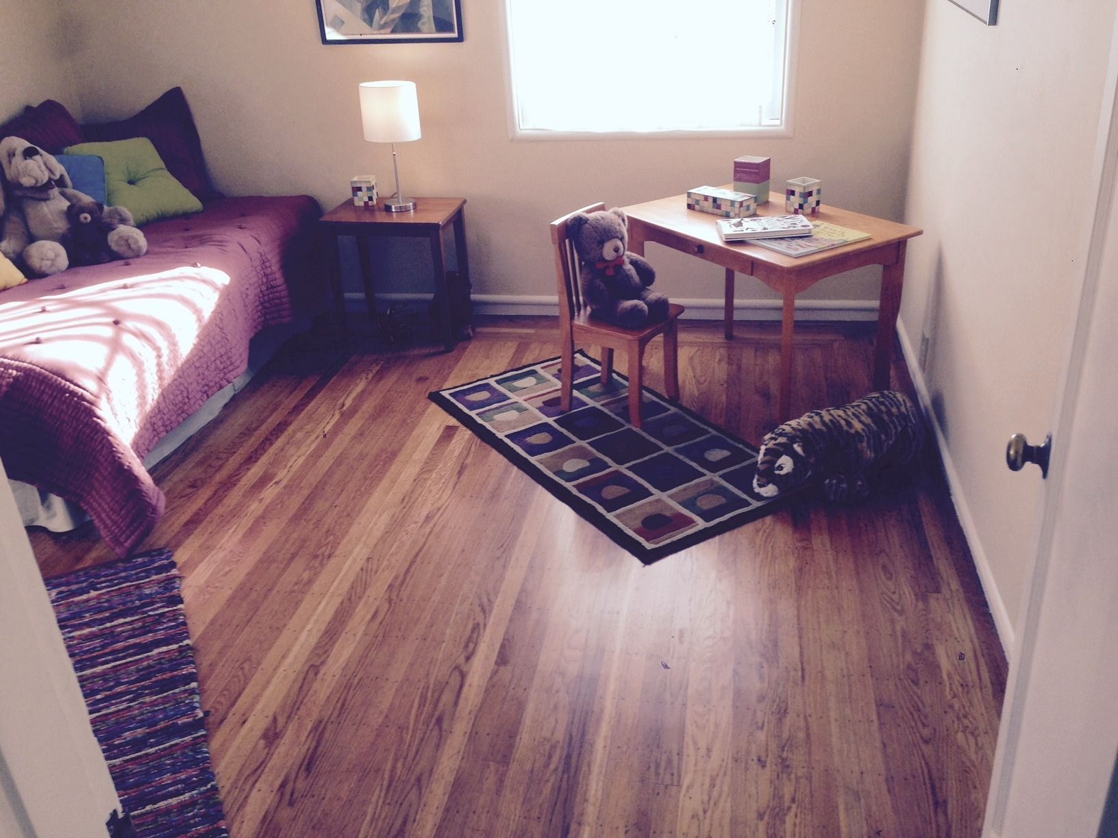 Bedroom with wooden floors, bed with purple cover, desk, rug, teddy bears, and natural light from a window.