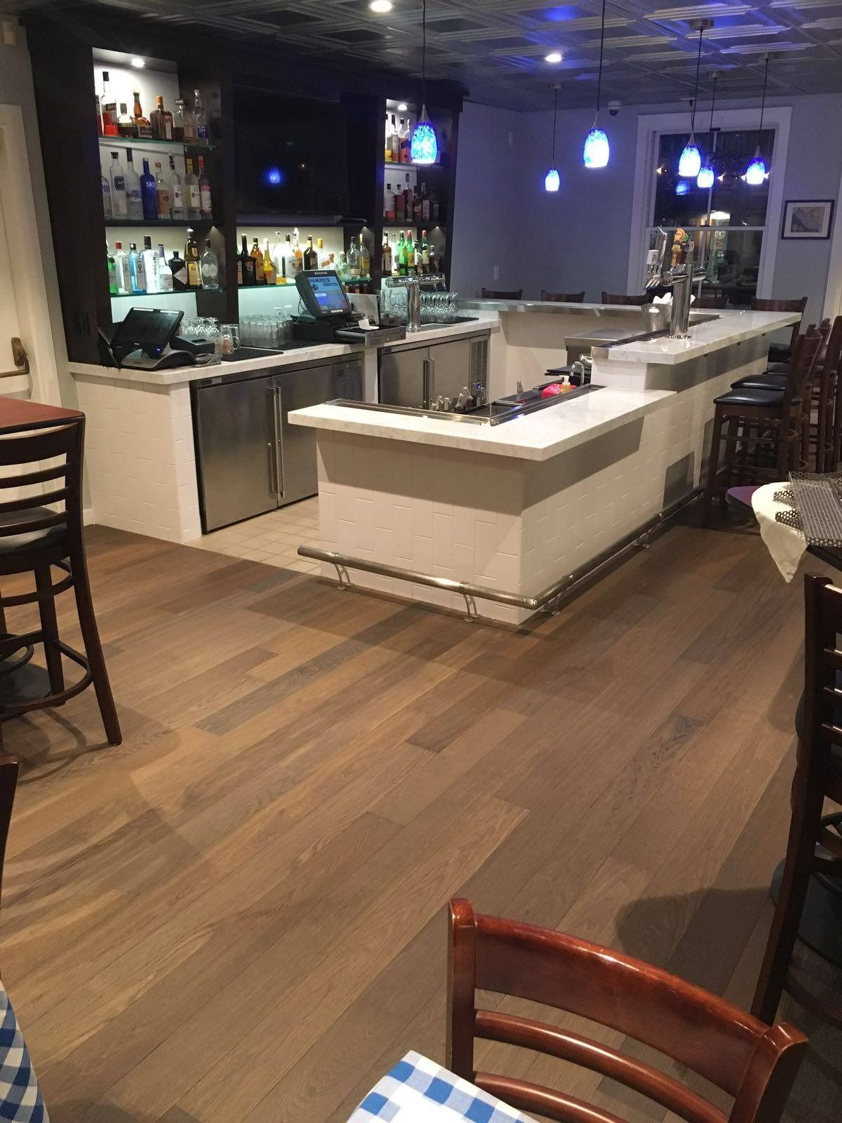 Bar interior with wood floors, white counter, shelves of liquor, and seating.