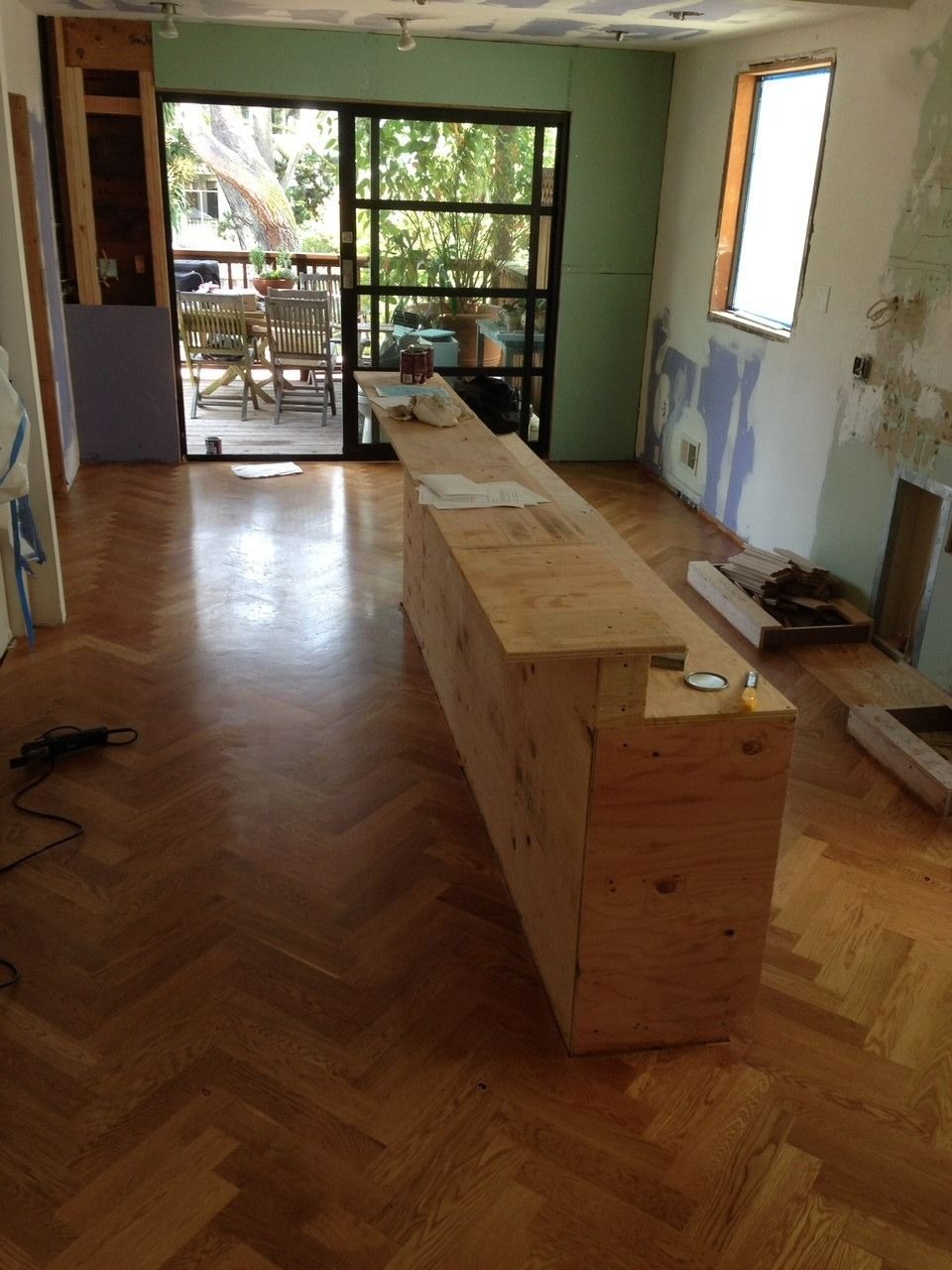 Room under renovation with a long, unfinished wooden structure on parquet flooring. Sliding glass door leads to a balcony.