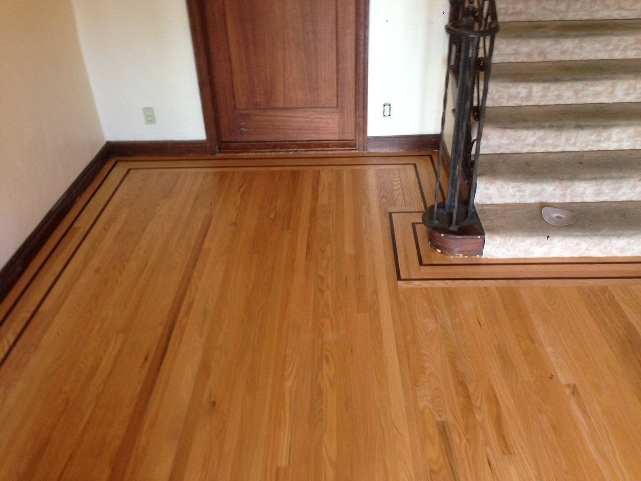 Wooden floor with a decorative border, next to a staircase and a wooden door.