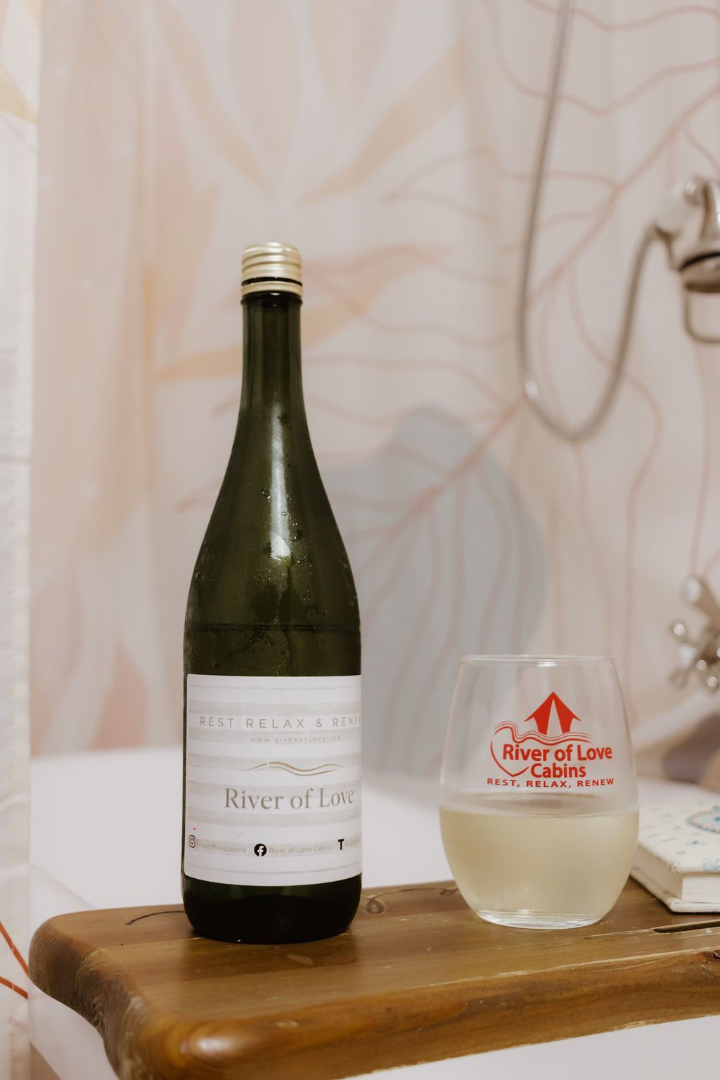 Bottle and glass of pale drink on a wooden table against a patterned curtain.