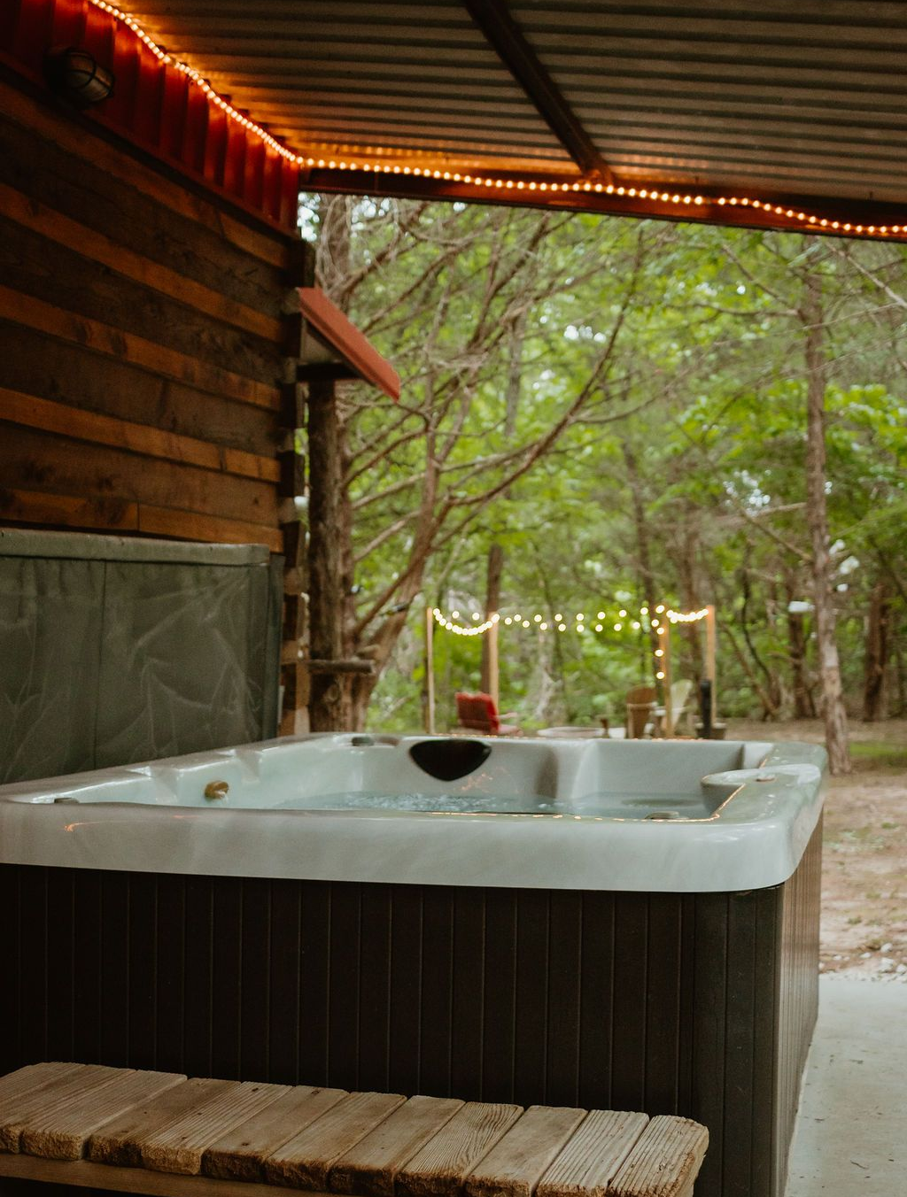 Hot tub on a covered wooden porch beside a wooded campsite, with string lights and trees beyond.