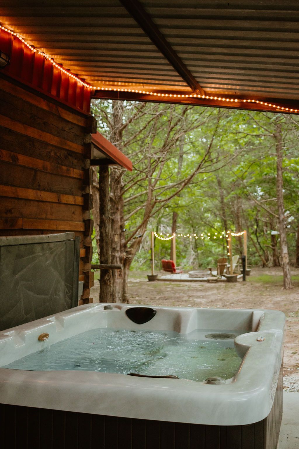 Covered hot tub under a rustic patio overlooking a wooded garden seating area