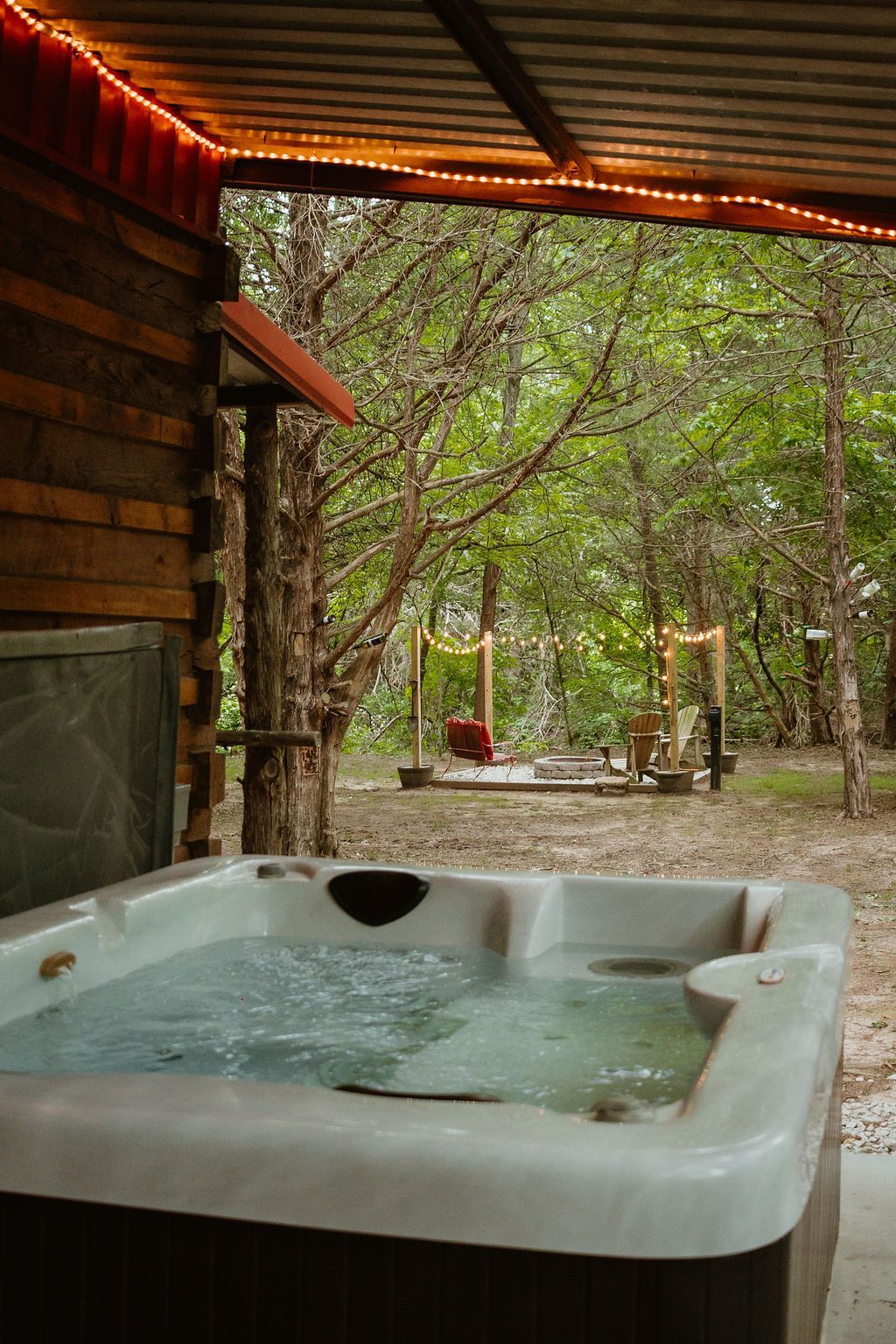 Outdoor hot tub beside a rustic cabin, with green forest and string lights in the background