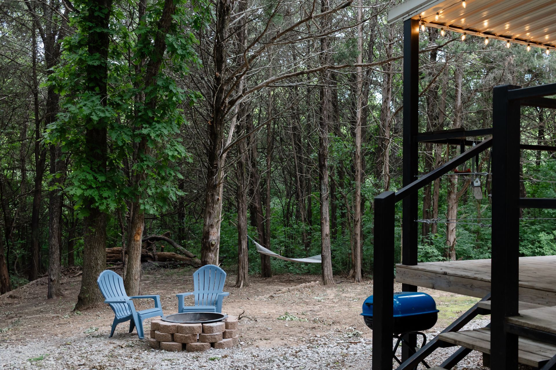 Two blue chairs around a fire pit in a wooded backyard beside a deck