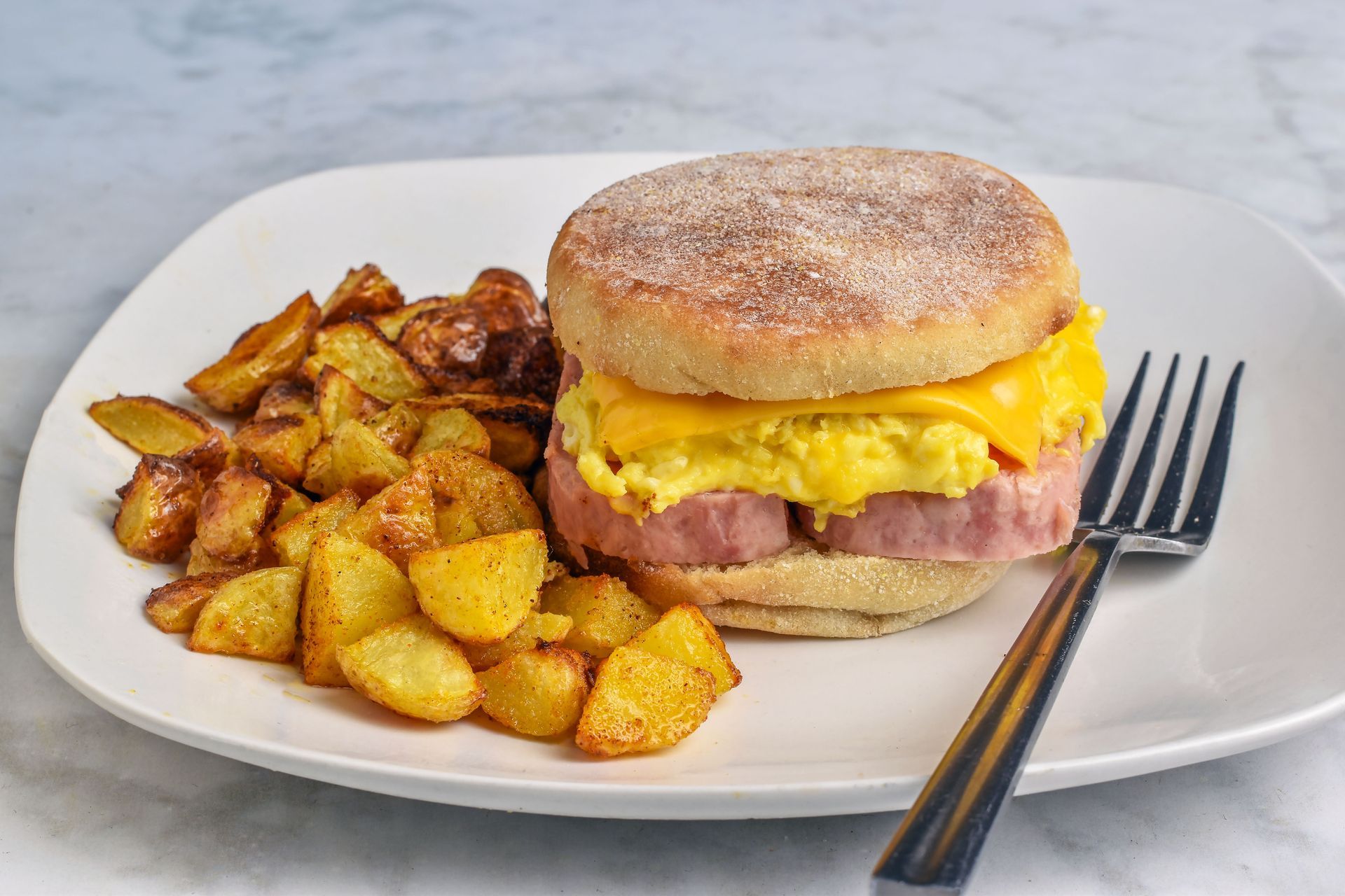 Breakfast sandwich with melted cheese and potatoes on a white plate with a fork