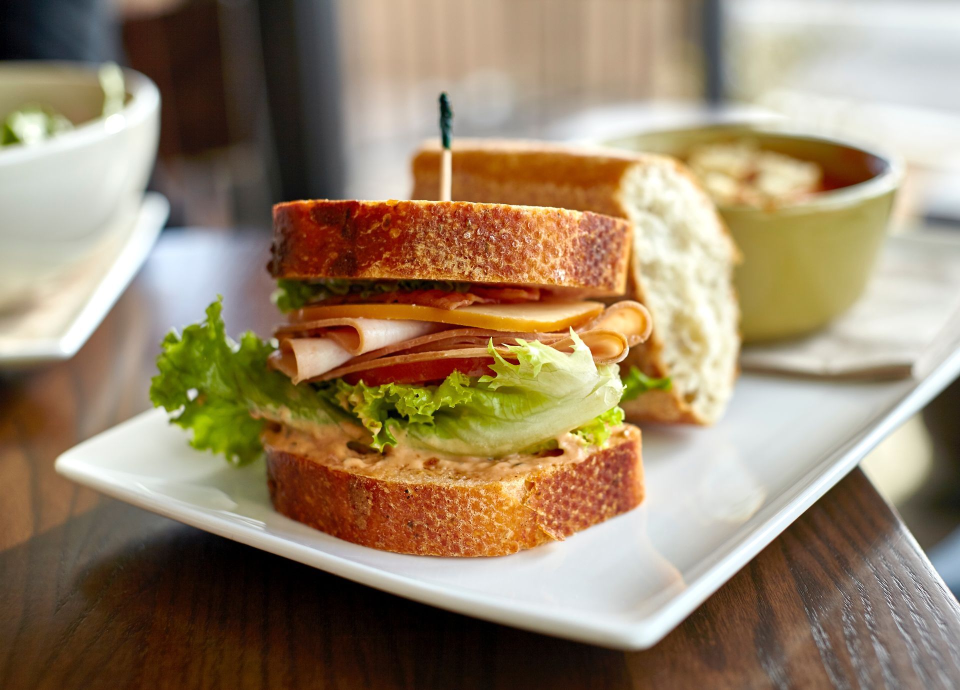 Sandwich on a white plate with lettuce and turkey, served with soup on a wooden table.