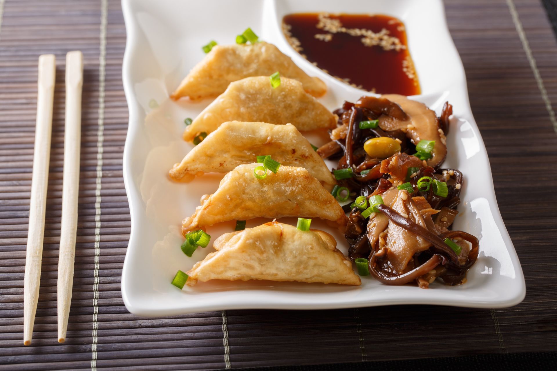 Potstickers with dipping sauce and sautéed mushrooms on a white platter, chopsticks beside them.