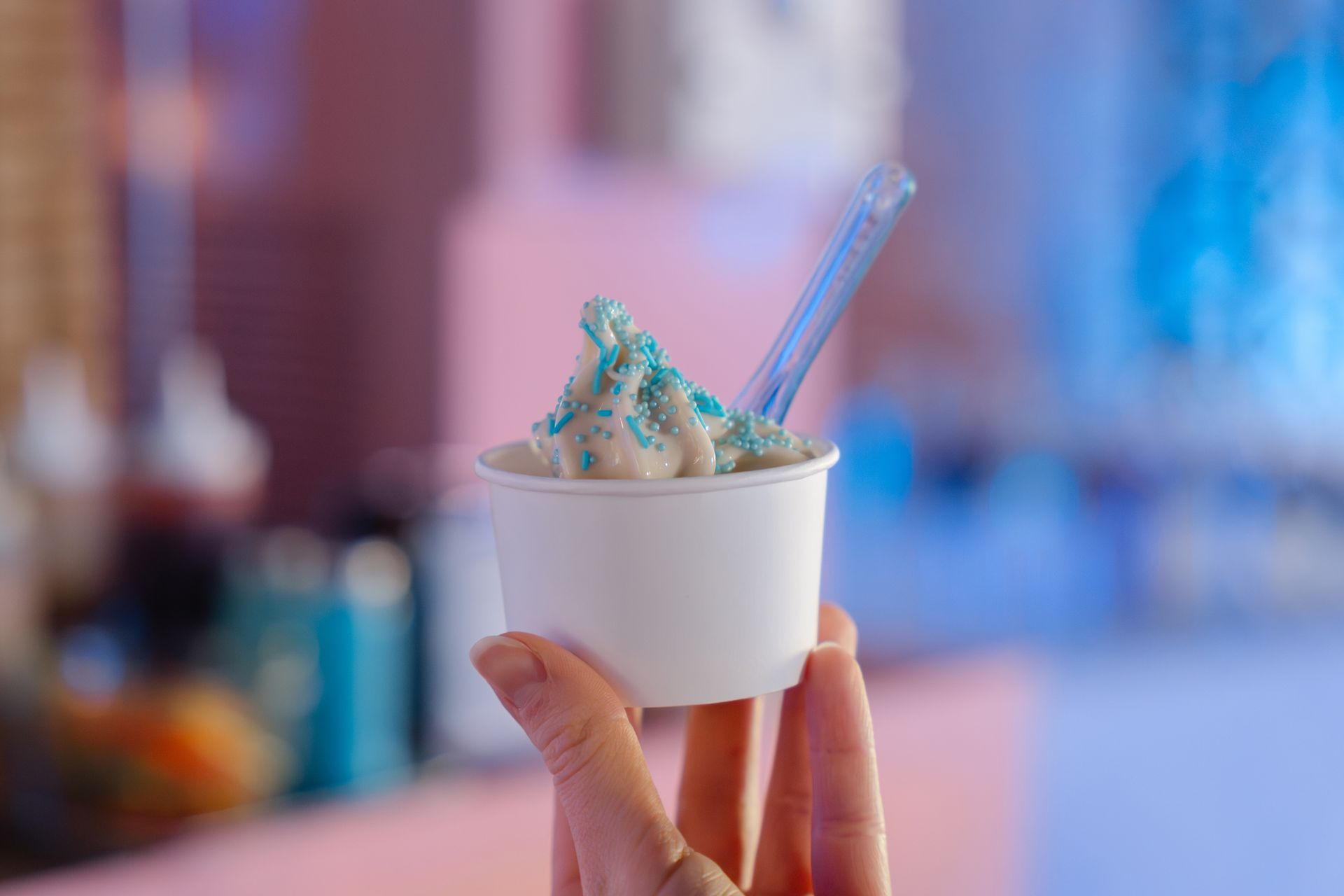 Hand holding a white cup of blue-topped soft serve with a spoon against a blurred colorful background