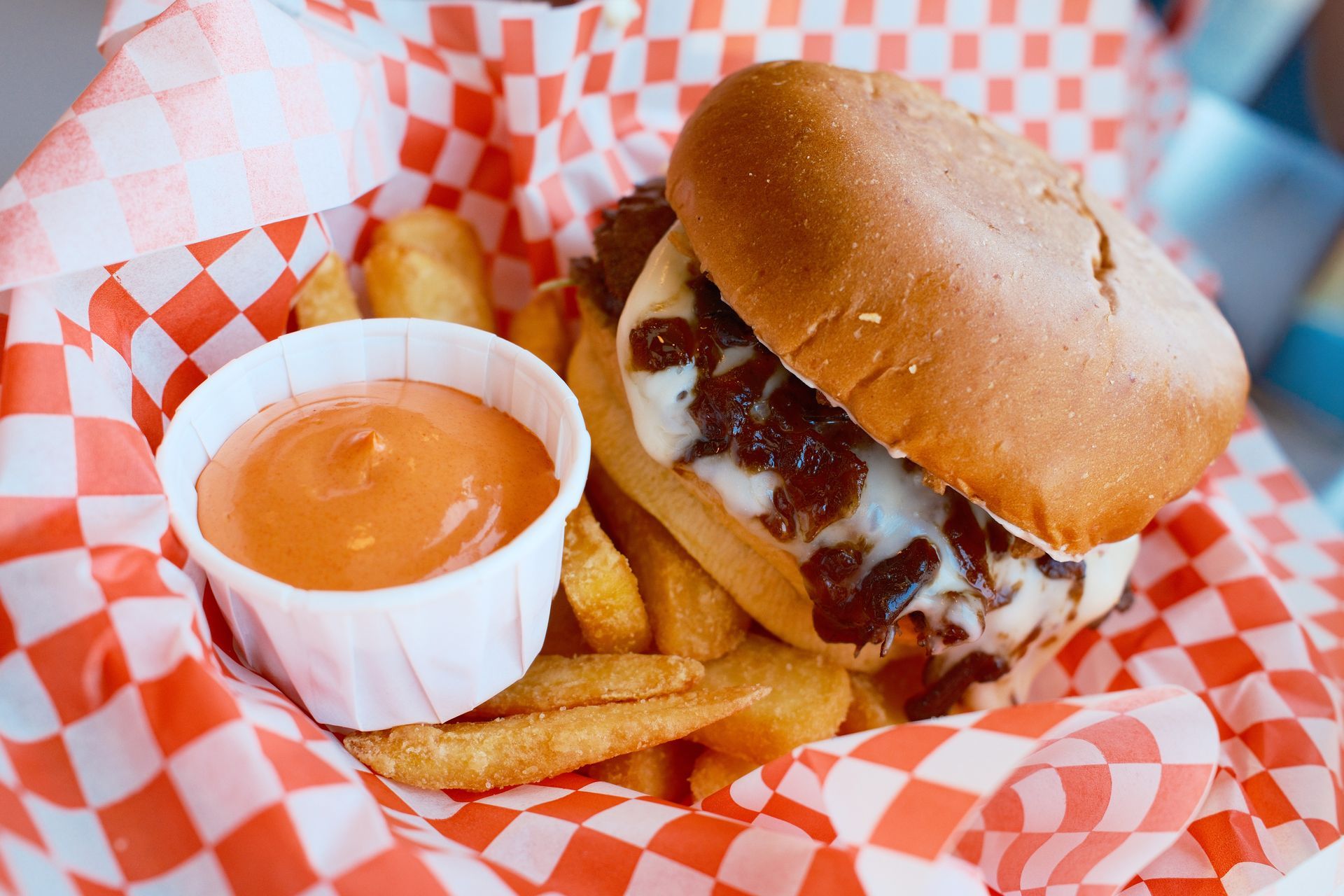 Burger and fries in a red-check paper basket with a side of orange dipping sauce