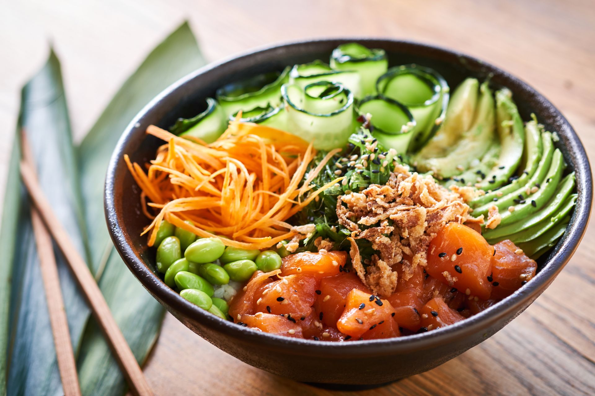 Bowl of colorful poke with avocado, cucumber, carrots, edamame, and salmon on a wooden table