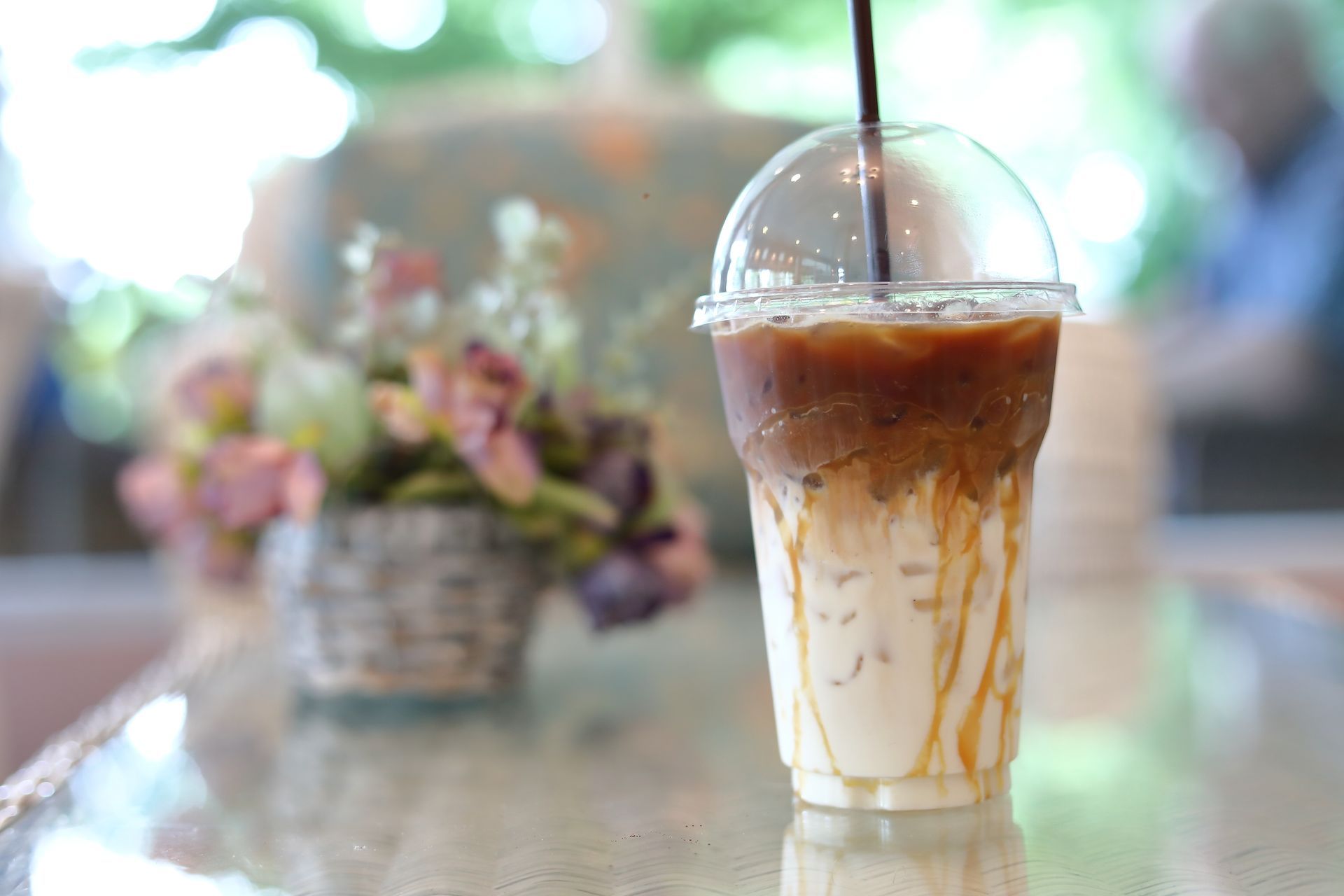 Iced coffee in a clear cup with a straw on a table, with a blurred flower arrangement in the background